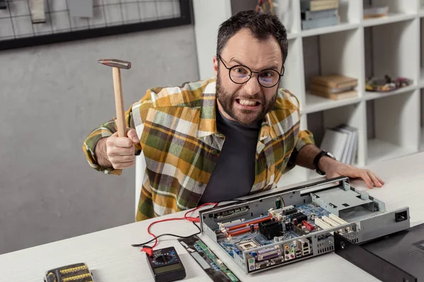 Reparador enojado con martillo en la mano contra la PC rota - foto de stock