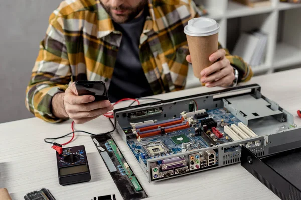 Imagen recortada del hombre con el café en la mano usando el teléfono inteligente sobre la PC rota - foto de stock