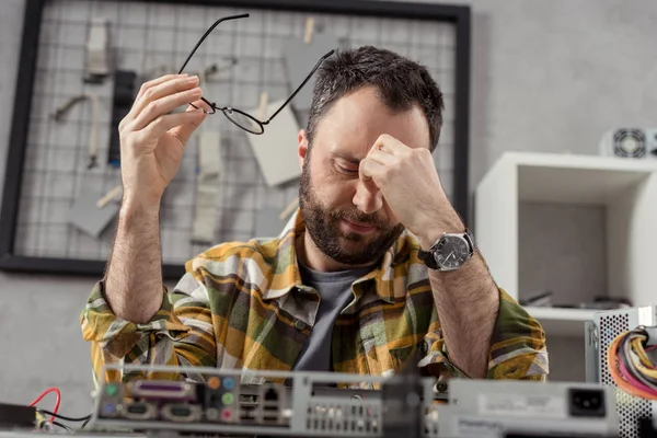 Hombre cansado sosteniendo gafas en la mano con los ojos cerrados contra el ordenador roto - foto de stock