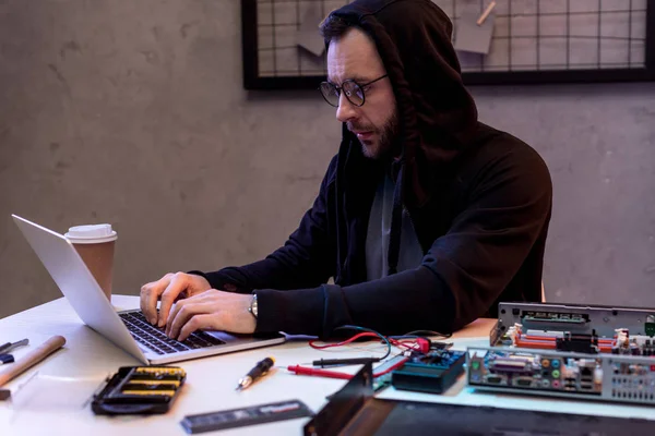 Hombre en sudadera con capucha usando el ordenador portátil cerca de PC roto - foto de stock