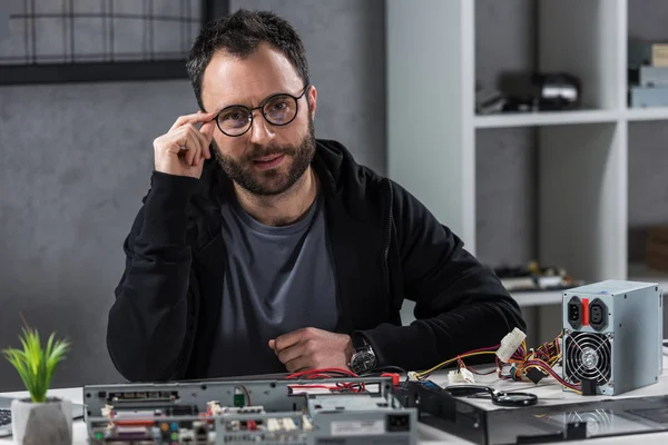 Mann mit Hand auf Brille blickt Kamera gegen Computerdetails auf Tisch — Stockfoto