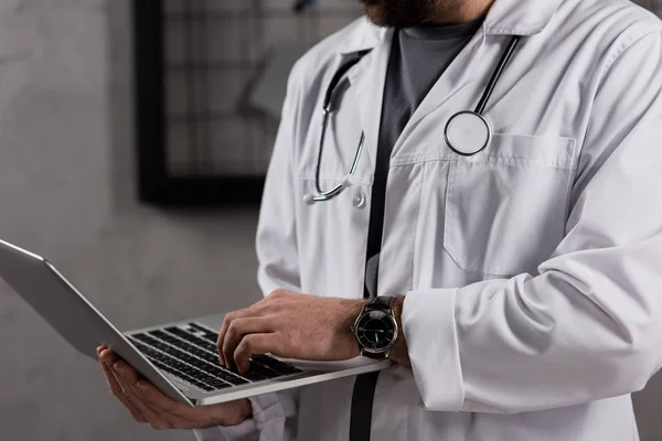 Image recadrée du médecin en manteau blanc avec stéthoscope sur le cou à l'aide d'un ordinateur portable — Photo de stock