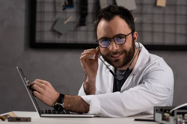 Médico sonriente con bata blanca y estetoscopio usando portátil mientras mira la cámara - foto de stock