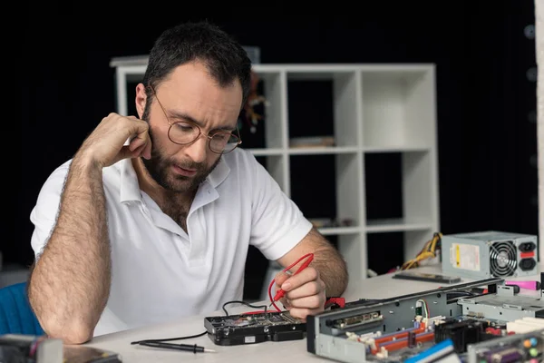 Repairman with hand on cheek using multimeter while testing hard disk drive — Stock Photo