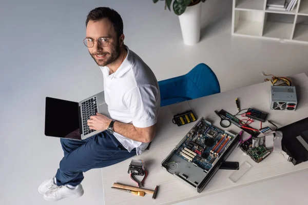 Hombre sonriente sentado en la mesa con portátil en las rodillas - foto de stock