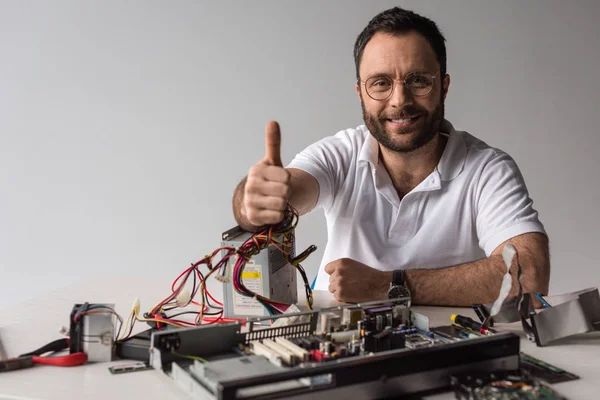 Hombre mostrando el pulgar hacia arriba mientras sonríe a la cámara contra la PC rota - foto de stock