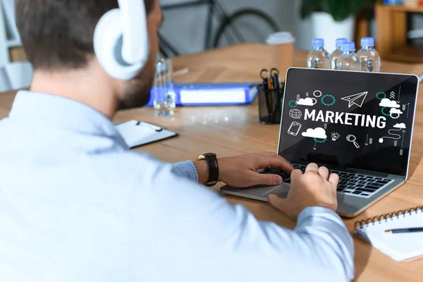 Abgeschnittene Aufnahme eines Geschäftsmannes mit Kopfhörern, der Musik hört und am Laptop arbeitet, mit Marketing-Wort und Symbolen auf dem Bildschirm — Stockfoto