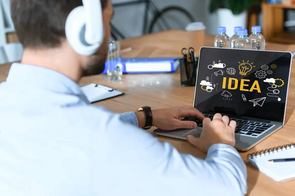 Recortado disparo de hombre de negocios en auriculares escuchando música y trabajando en el ordenador portátil con la palabra idea y los iconos en la pantalla - foto de stock