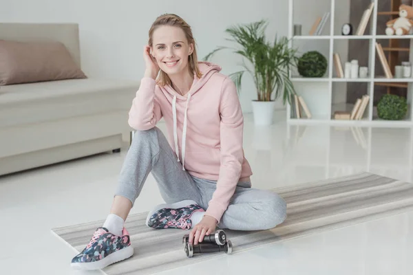 Jolie femme assise sur un tapis de yoga avec haltères à la maison — Photo de stock