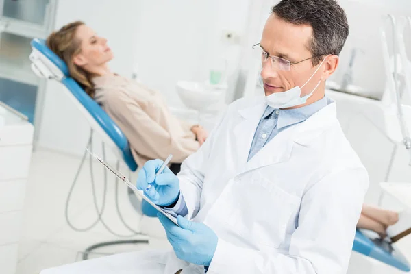 Dentista gravando diagnóstico enquanto paciente do sexo feminino esperando na clínica moderna — Fotografia de Stock