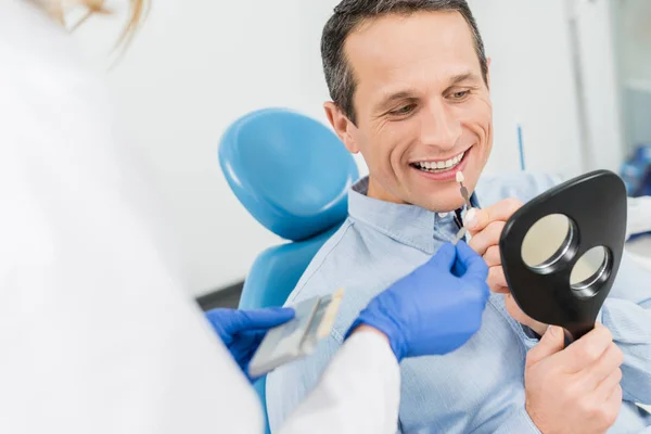 Paciente masculino escolhendo implante dentário olhando para o espelho na clínica odontológica moderna — Fotografia de Stock