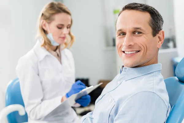 Dentist recording diagnosis while male patient waiting in modern clinic — Stock Photo