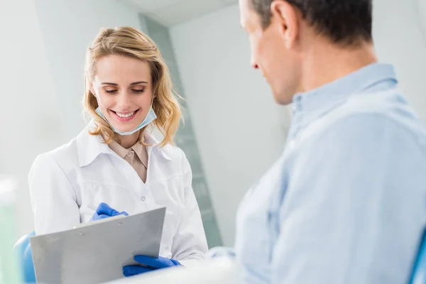 Médico paciente y sonriente registrando diagnóstico en clínica dental moderna - foto de stock