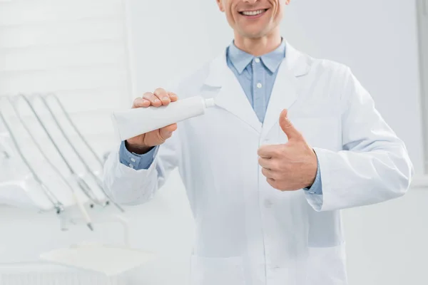 Visão de perto do médico mostrando tubo de pasta de dentes em branco e polegar para cima na clínica odontológica moderna — Fotografia de Stock