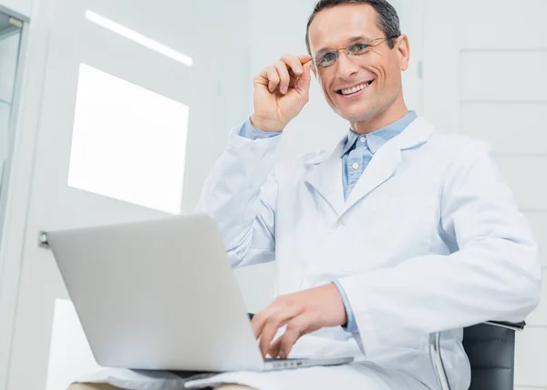 Усміхнений лікар працює ноутбуком у сучасній клініці — стокове фото
