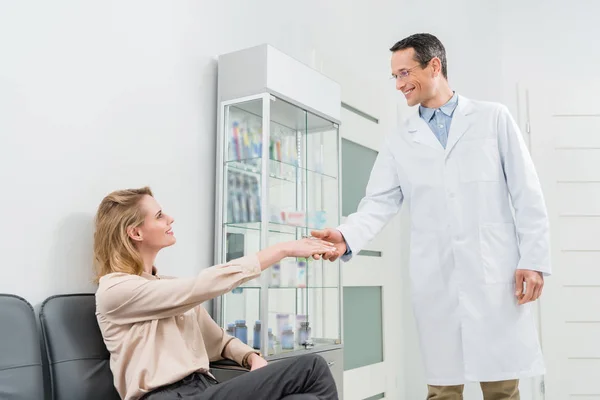 Médico sonriente saludando paciente mujer estrechando las manos en clínica dental moderna - foto de stock