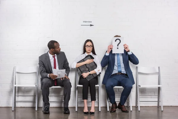 Empresário cobrindo cara com cartão com ponto de interrogação enquanto espera por entrevista de emprego com pessoas de negócios multiétnicos — Fotografia de Stock
