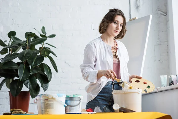 Young inspired girl holding palette and looking at camera in light studio — Stock Photo