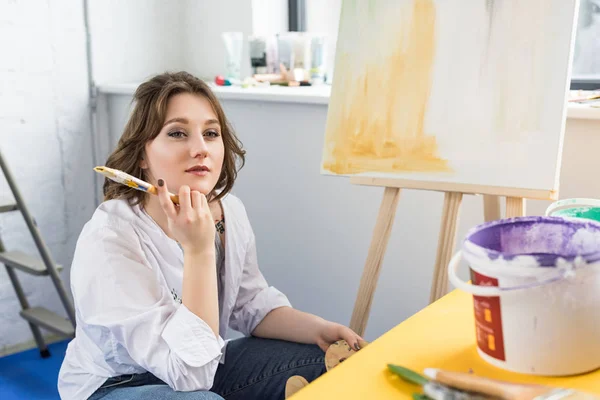 Young creative girl dreaming at easel in light studio — Stock Photo