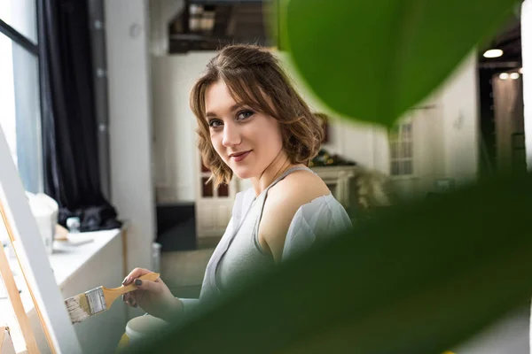 Joven chica artística mirando a la cámara a través de hojas mientras pinta en estudio de luz - foto de stock