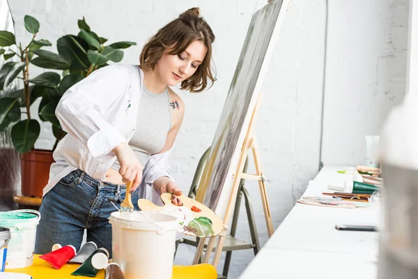 Young inspired girl dunks a paintbrush into the paint in light studio — Stock Photo