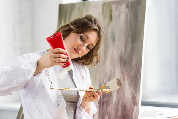 Young creative girl squeezing paint tube in light studio — Stock Photo