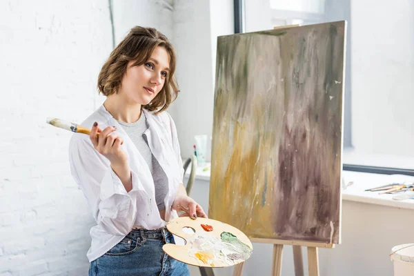 Joven chica inspirada soñando con la paleta en la mano en el estudio de luz - foto de stock