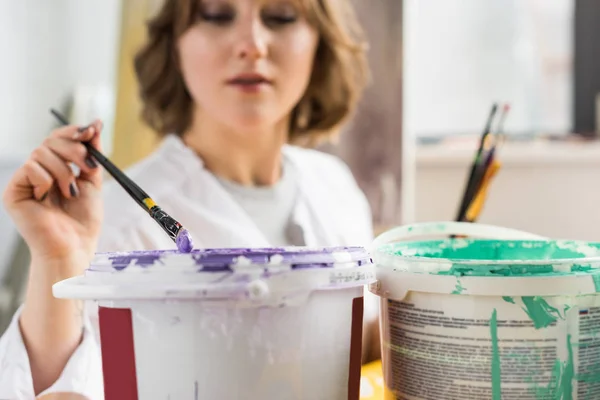 Young creative girl dunks a paintbrush into the paint in light studio — Stock Photo