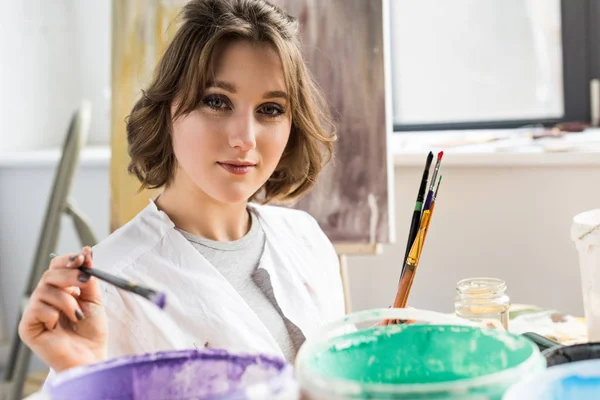 Young artistic girl dunks a paintbrush into the paint in light studio — Stock Photo