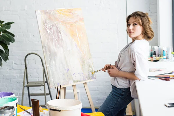 Young artistic girl standing by easel with brush in light studio — Stock Photo