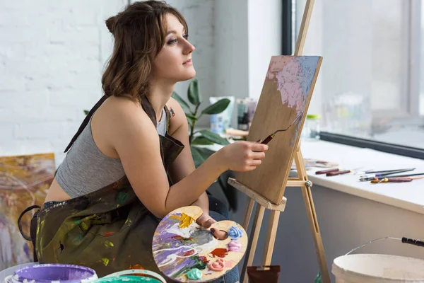 Joven chica creativa soñando por caballete en estudio de luz - foto de stock