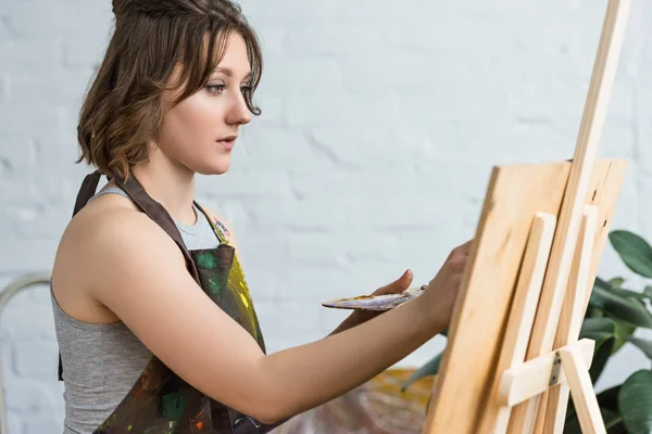Young inspired girl working by easel in light studio — Stock Photo