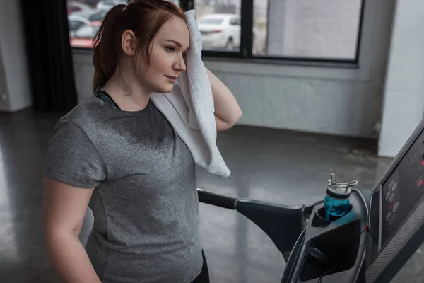 Chica con sobrepeso limpiándose la cara mientras corre en la cinta de correr en el gimnasio - foto de stock