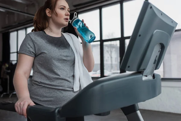 Курчавая девушка пьет из бутылки с водой во время бега на беговой дорожке в спортзале — стоковое фото
