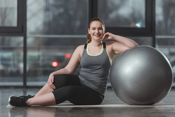 Ragazza in sovrappeso seduta sul pavimento con palla fitness in palestra — Foto stock