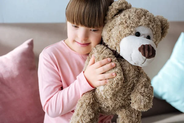 Kind mit Down-Syndrom sitzt auf Sofa und umarmt Teddybär — Stockfoto