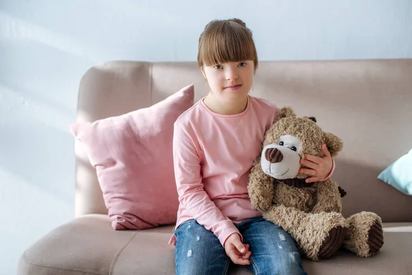 Niño con síndrome de Down sentado en el sofá y abrazando oso de peluche - foto de stock