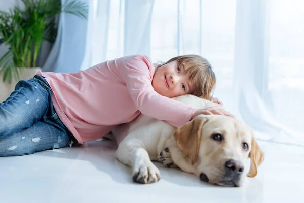 Glückliches Kind mit Down-Syndrom und Labrador Retriever kuschelt auf dem Boden — Stockfoto