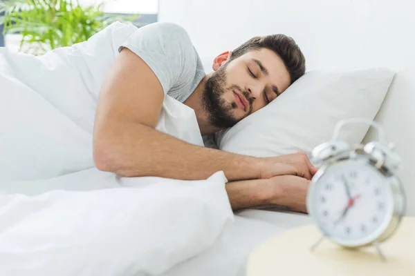 Красивый бородатый мужчина спит в постели по утрам, будильник на переднем плане — стоковое фото