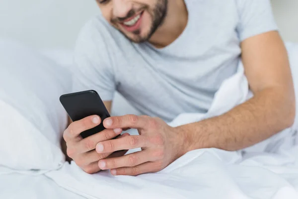 Homme barbu souriant couché sur le lit et utilisant un smartphone — Photo de stock