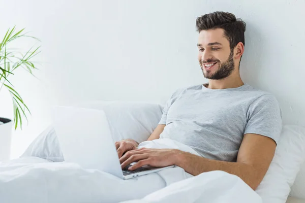 Smiling man lying on bed and using laptop — Stock Photo