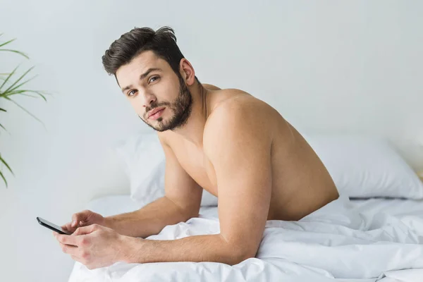 Schöner hemdloser Mann, der morgens im Bett auf dem Smartphone Nachrichten sendet — Stockfoto