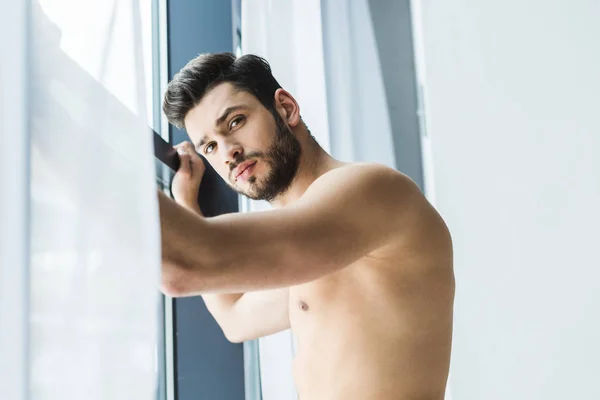 Hombre sin camisa de pie en la ventana y mirando a la cámara - foto de stock