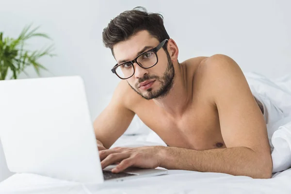 Hombre guapo en gafas usando el ordenador portátil en la cama por la mañana - foto de stock