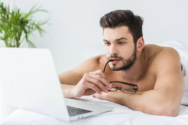 Thoughtful man with eyeglasses using laptop in bed in the morning — Stock Photo