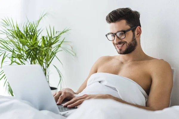 Schöner lächelnder Mann mit Laptop im Bett — Stockfoto