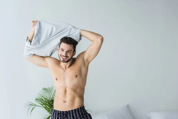 Handsome sexy smiling man taking off his t-shirt in bedroom — Stock Photo