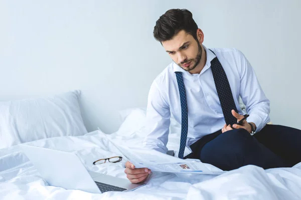 Schöner Geschäftsmann liest Zeitung auf dem Bett mit Laptop — Stockfoto