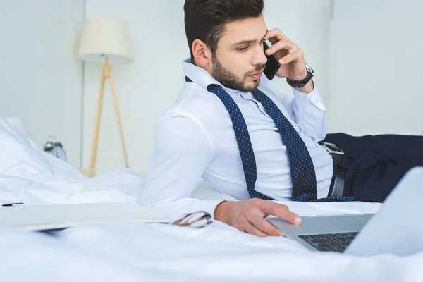 Hombre de negocios guapo acostado en la cama mientras trabaja con el ordenador portátil y el teléfono inteligente - foto de stock