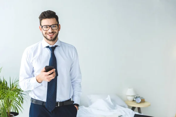Smiling businessman in eyeglasses using smartphone in bedroom — Stock Photo